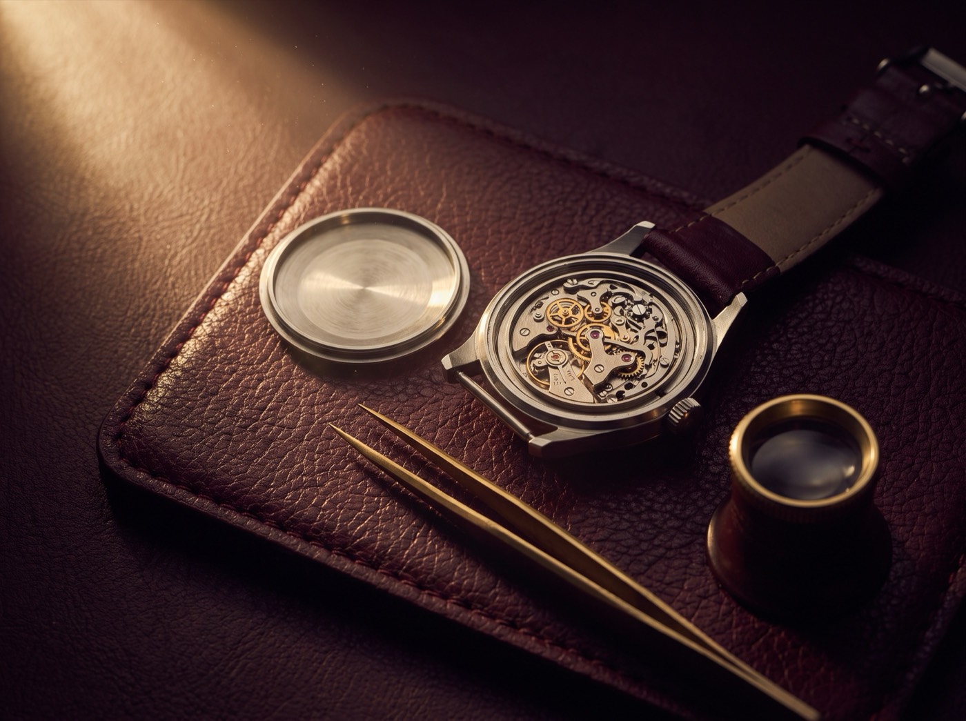 A wristwatch with its caseback open beside brass tweezers and a jeweler's loupe on an oxblood leather bench pad